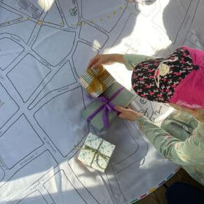 Edukacinis užsiėmimas darželinukams apie Vilniaus istoriją Mergaitė edukacijoje vaikams apie Vilnių tyrinėja didelį žemėlapį ir supakuotas dovanas, kuriose daiktai susiję su Vilniaus istorija.